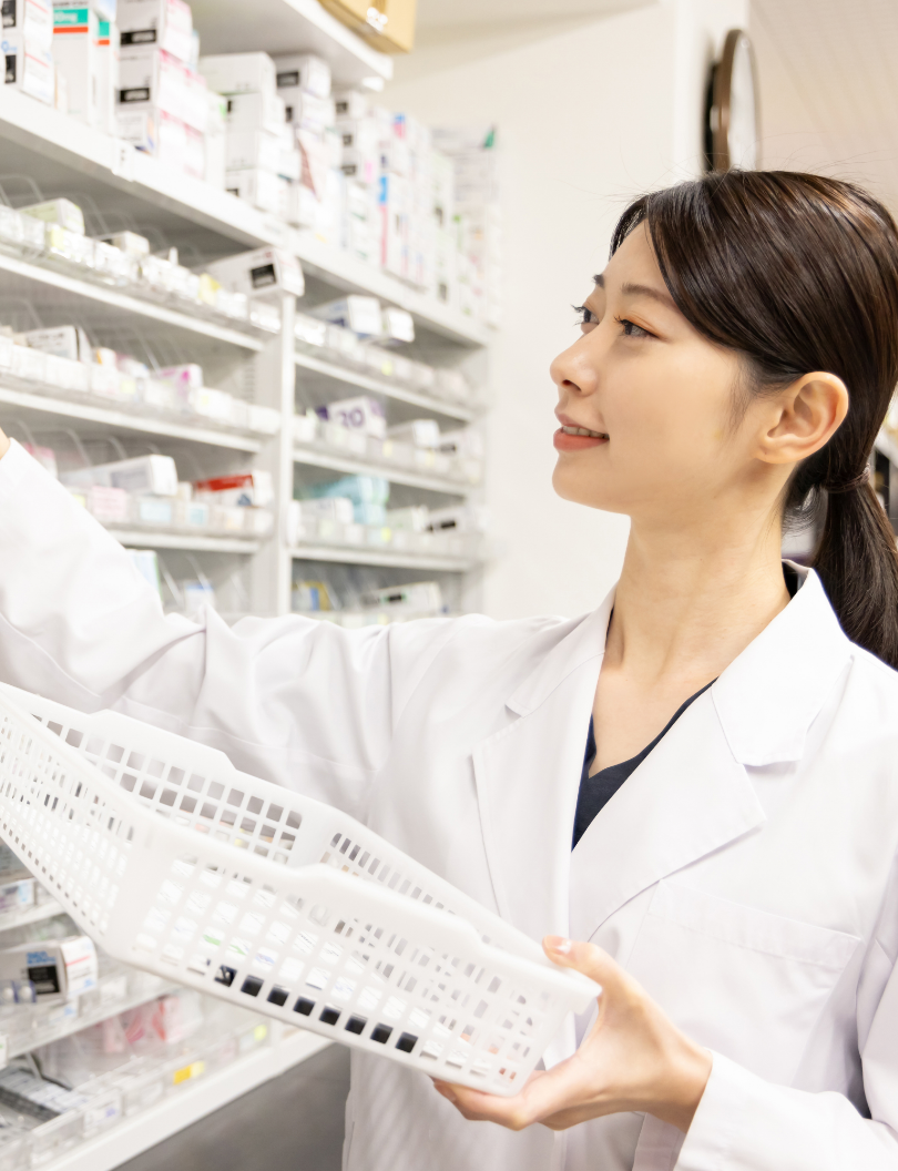 白衣を着た薬剤師が、調剤室の棚から薬を取り出しバスケットに入れている様子